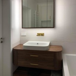 Egad - Nowoczesna łazienka z umywalką nablatową na drewnianej szafce z szufladami i złotymi uchwytami, lustro w brązowej ramie, białe płytki z delikatną fakturą na ścianie, widoczna część wanny.