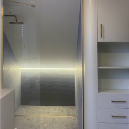 Egad - Nowoczesna łazienka na poddaszu z szarą mozaiką, złotymi akcentami, przeszkloną kabiną prysznicową i zabudową meblową z szufladami.