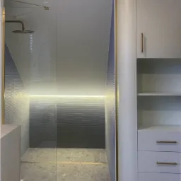 Nowoczesna łazienka na poddaszu z szarą mozaiką, złotymi akcentami, przeszkloną kabiną prysznicową i zabudową meblową z szufladami.