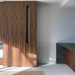 Nowoczesna kuchnia z zabudową z forniru orzechowego, minimalistyczny design, pionowy uchwyt w szafce, blat kuchenny z szarego kamienia, białe ściany i podłoga, oświetlenie słoneczne.