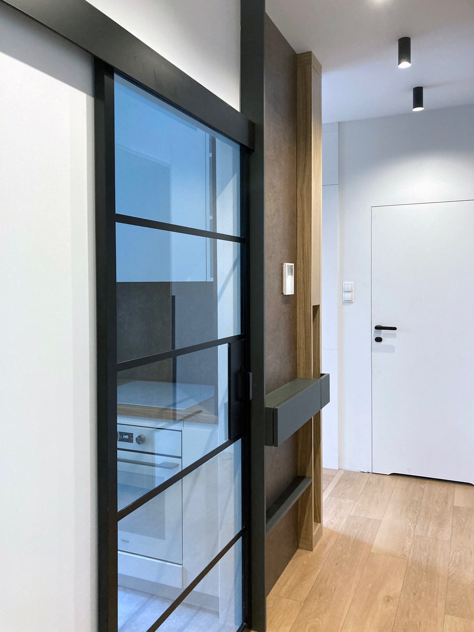 Przeszklone, przesuwne drzwi w czarnej ramie oddzielające korytarz od kuchni, widok na białą zabudowę kuchenną i piekarnik TEKA, drewniana podłoga i minimalistyczne oświetlenie.