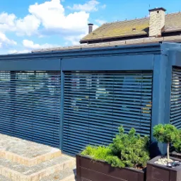 Szare żaluzje fasadowe poziome, osłaniające taras z roślinami w donicach. Widok z zewnątrz, w tle budynek z cegły i błękitne niebo.