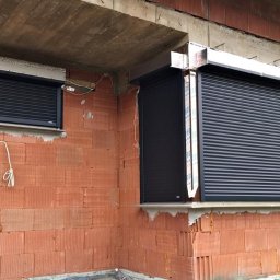 ALUPLASTI S.C. MAGDALENA I PIOTR GAZDA - Ciemne rolety zamontowane w oknach budynku w trakcie budowy, widoczne cegły i surowy beton. Roleta po prawej stronie z listwami ochronnymi na boku.