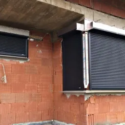 Ciemne rolety zamontowane w oknach budynku w trakcie budowy, widoczne cegły i surowy beton. Roleta po prawej stronie z listwami ochronnymi na boku.