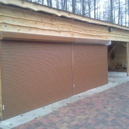 ALUPLASTI S.C. MAGDALENA I PIOTR GAZDA - Brązowa roleta na drewnianym garażu z dobudowaną wiatą, widok na kostkę brukową i drzewa w tle. Zamknięta, pozioma roleta na garażu z desek.
