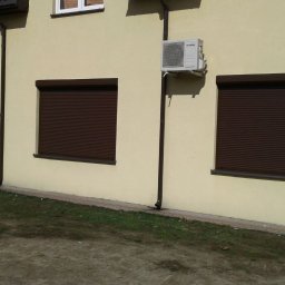 ALUPLASTI S.C. MAGDALENA I PIOTR GAZDA - Brązowe rolety zewnętrzne na elewacji budynku z kremową fasadą. Widoczne trzy okna z opuszczonymi roletami i klimatyzator na ścianie.