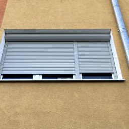 Aluroli - Zamknięta szara roleta zewnętrzna na oknie budynku o żółtej elewacji. Widoczna rynna. Detal architektoniczny, ochrona, nowoczesność.