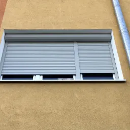 Zamknięta szara roleta zewnętrzna na oknie budynku o żółtej elewacji. Widoczna rynna. Detal architektoniczny, ochrona, nowoczesność.