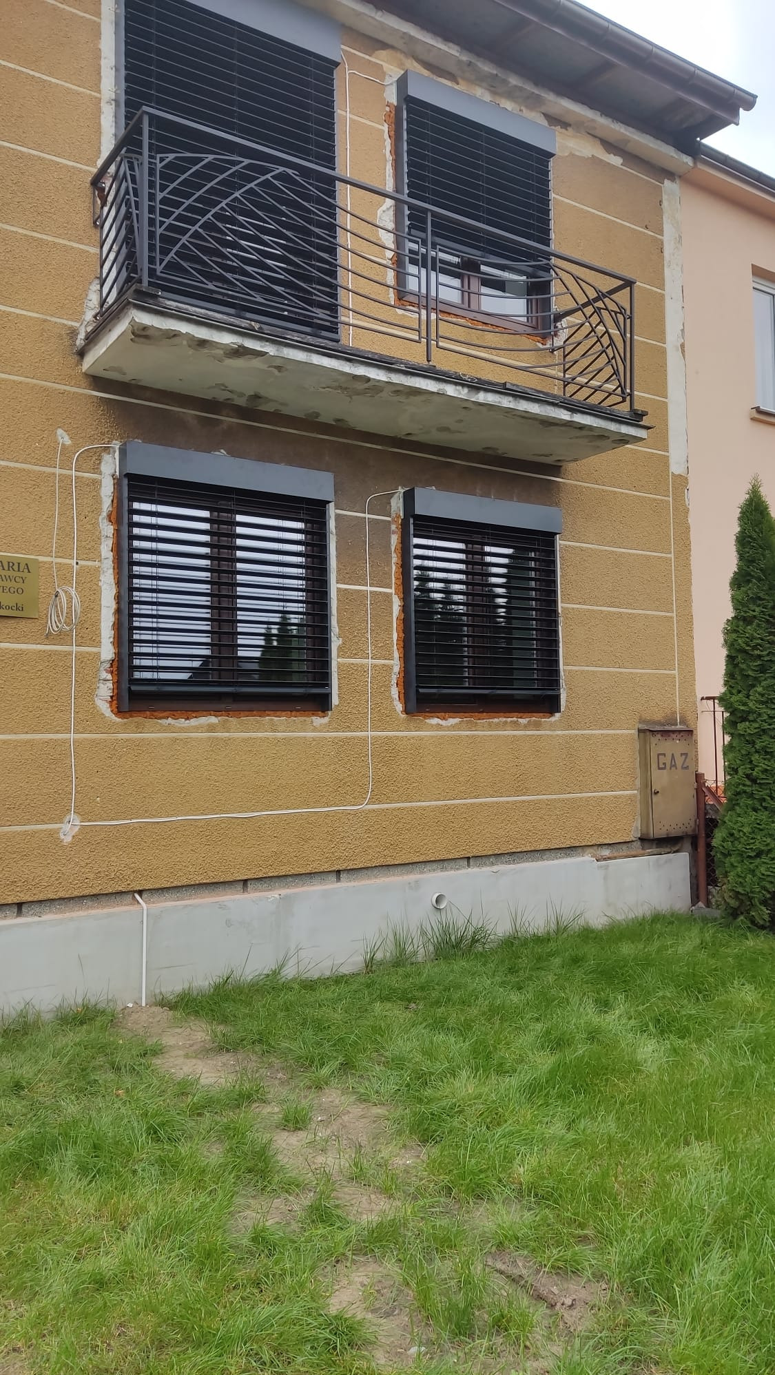 Elewacja budynku z dwoma oknami i balkonem, na których zamontowano czarne żaluzje fasadowe, z widoczną skrzynką gazową i fragmentem trawnika.