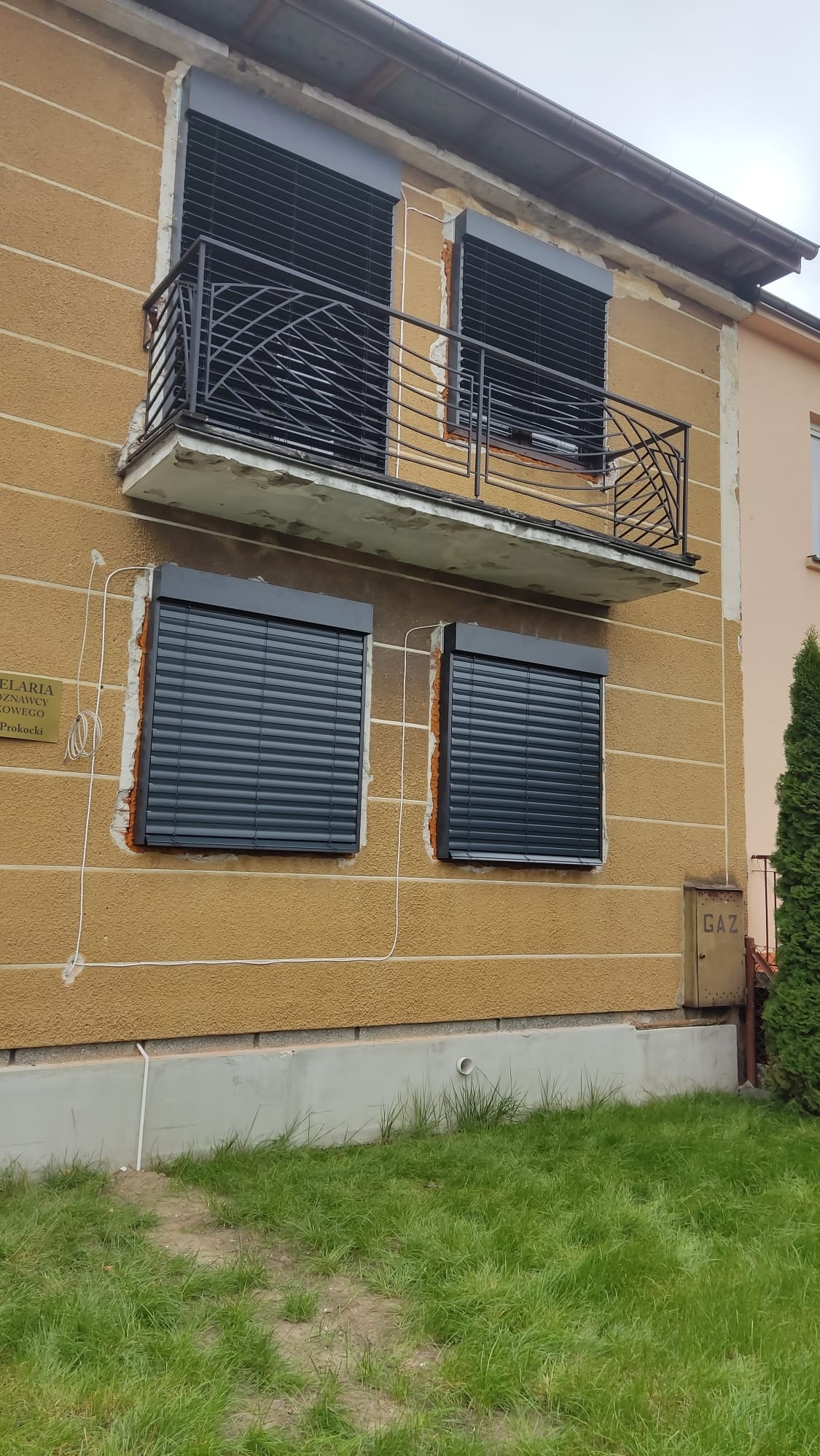 Zamontowane rolety antywłamaniowe w oknach budynku z balkonem. Widoczne ślady po montażu na elewacji. Dolne okna z roletami na parterze, górne na balkonie.