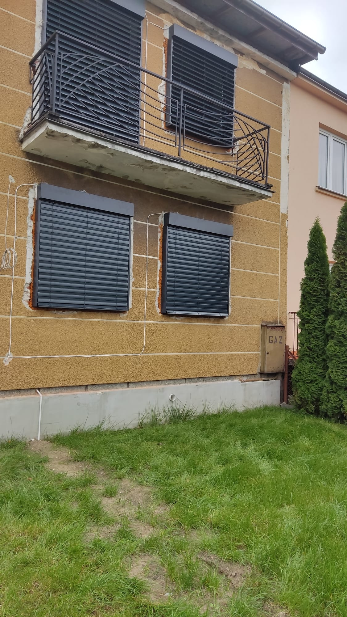 Elewacja domu z oknami zabezpieczonymi szarymi roletami. Na górze balkon z ozdobną, metalową balustradą. Widoczne ślady po montażu.