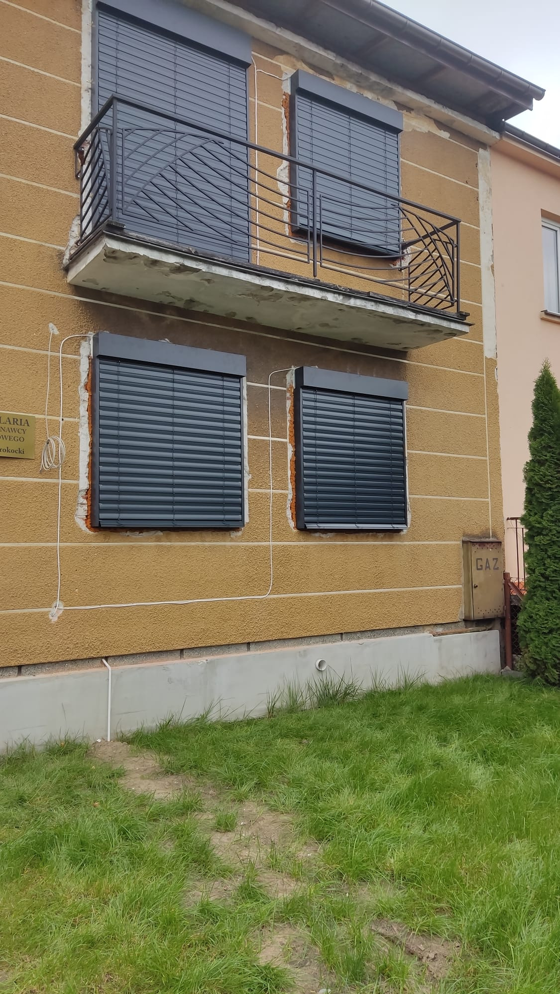 Dom z beżową elewacją i zamontowanymi, grafitowymi roletami zewnętrznymi. Na piętrze balkon z ozdobną balustradą. Widoczna skrzynka gazowa.
