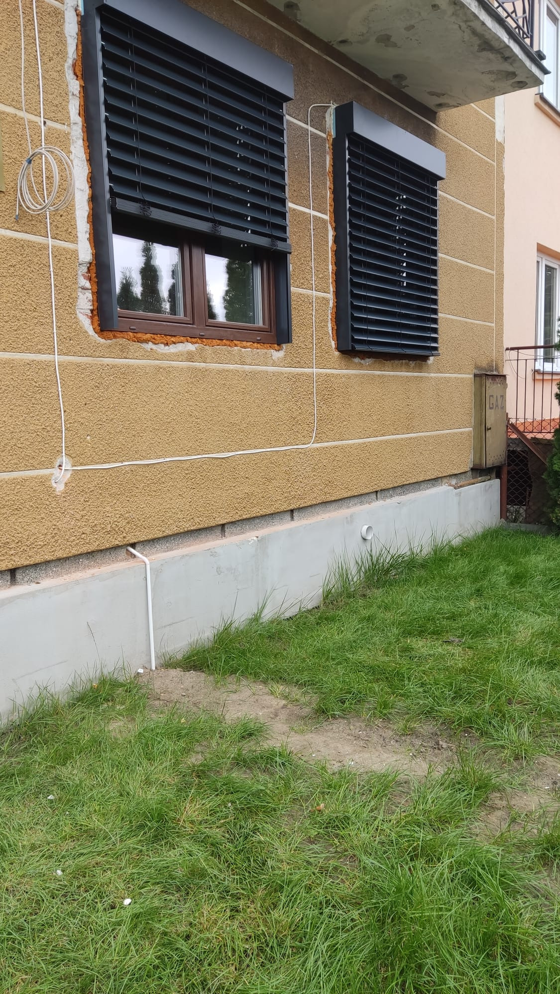 Ciemne żaluzje fasadowe zamontowane na oknach budynku z ociepleniem. Widoczne przewody na elewacji i trawnik przed budynkiem. Kąt ujęcia z dołu.