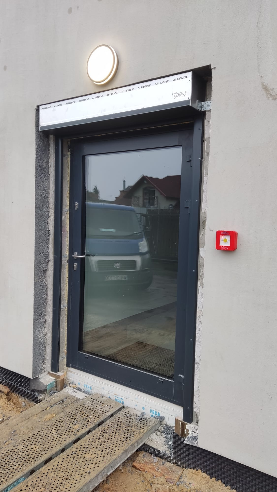 Montaż okna z roletą zewnętrzną w surowym stanie. Widoczne rusztowanie i lampa nad oknem. W szybie odbija się samochód dostawczy i budynek.