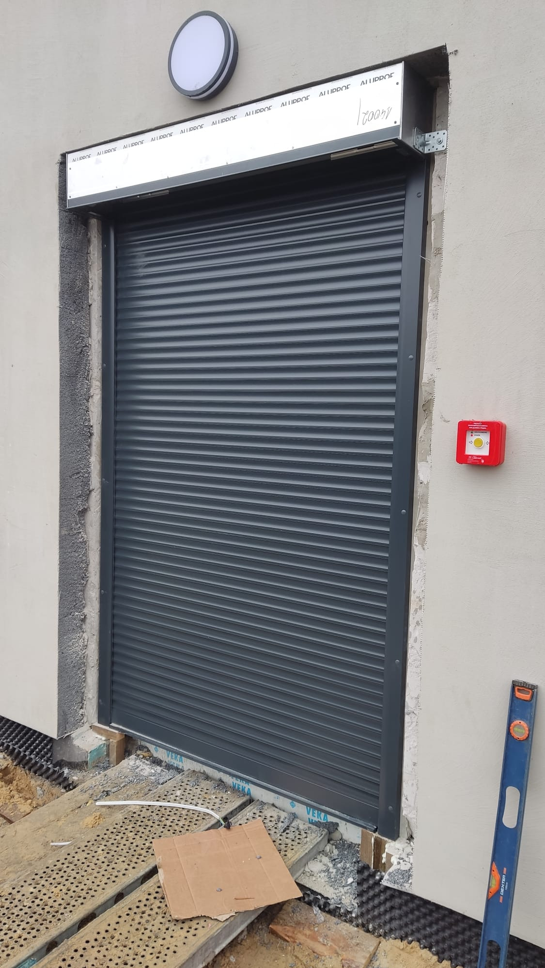 Zamknięta szara roleta zewnętrzna na tle surowej elewacji. Widoczny fragment rusztowania i poziomica. Na ścianie lampa i czerwony przycisk.