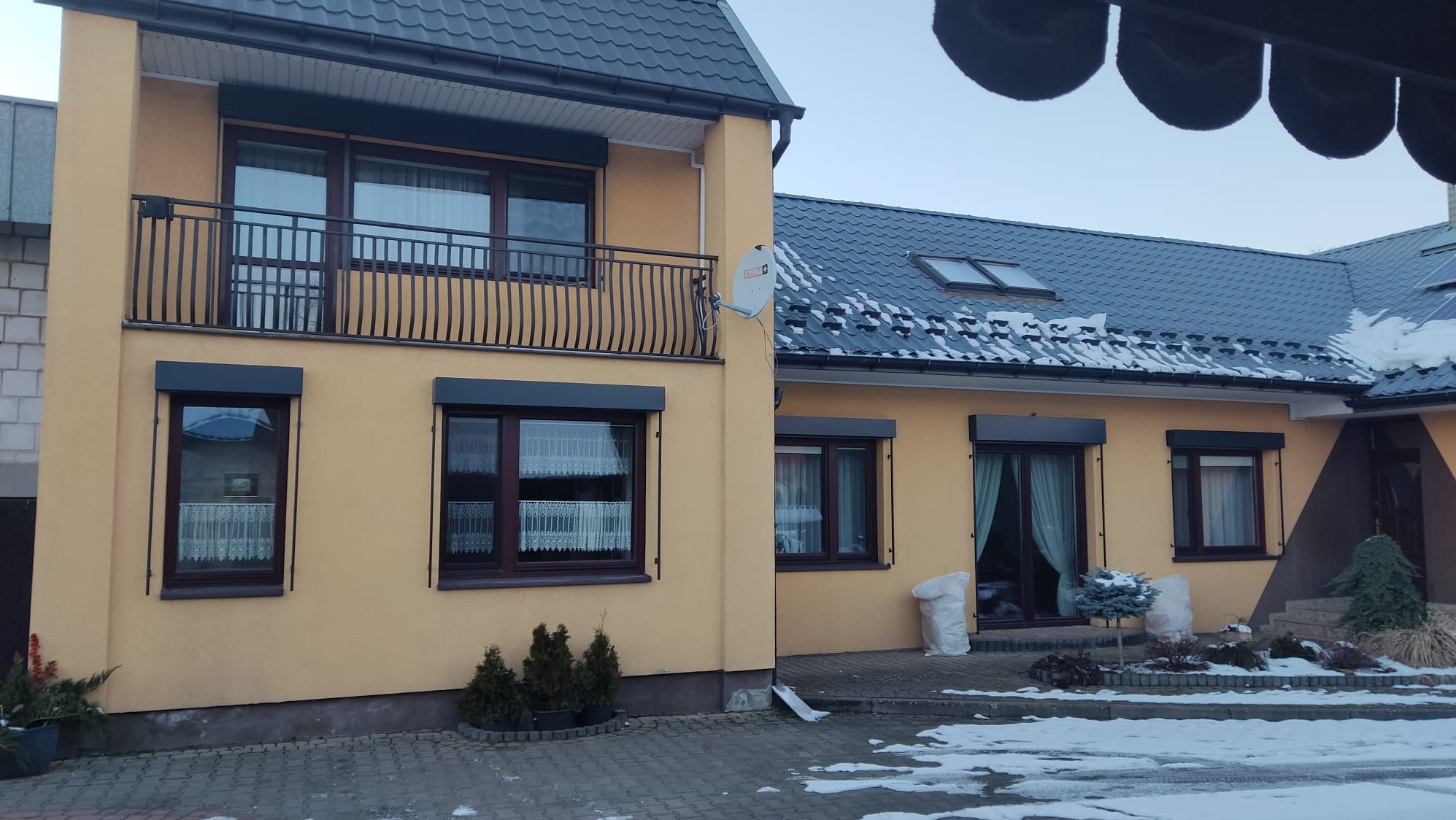 Żółty dom z brązowymi oknami i szarymi roletami zewnętrznymi, częściowo opuszczonymi. Widoczny balkon z balustradą i antena satelitarna na elewacji.