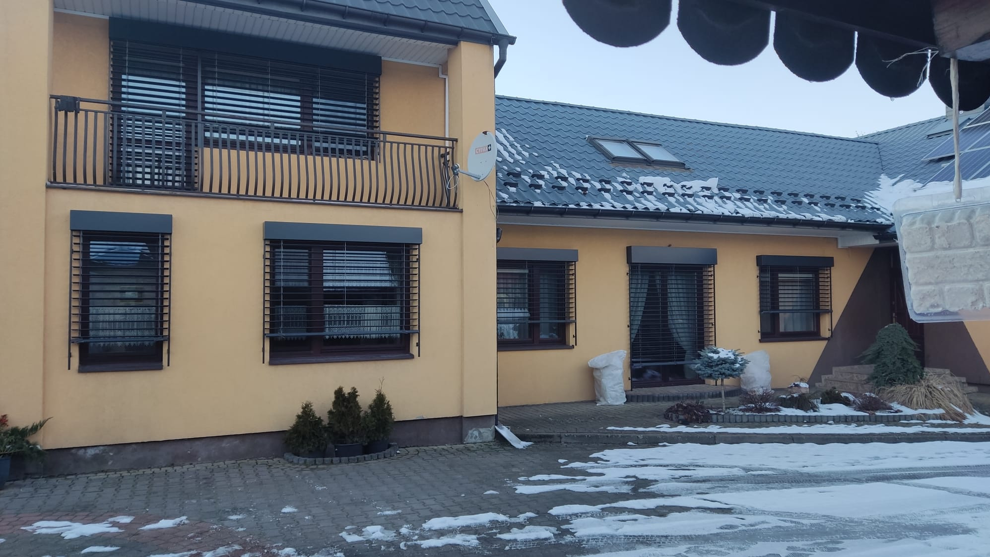 Dom z oknami zabezpieczonymi roletami antywłamaniowymi w kolorze antracytowym. Zima, śnieg na dachu i chodniku. Balustrada balkonowa z metaloplastyki.