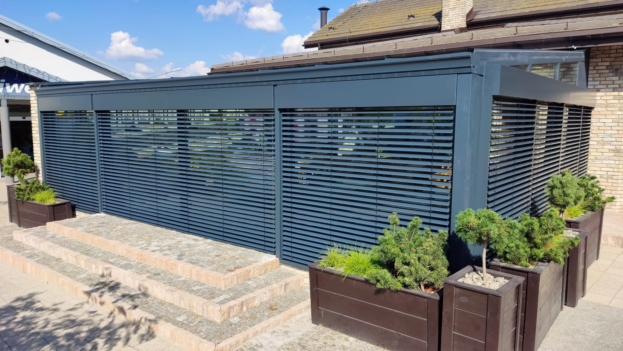 Nowoczesna, grafitowa zabudowa tarasu z poziomymi żaluzjami fasadowymi, uzupełniona o donice z zielenią. Widok na budynek w tle.