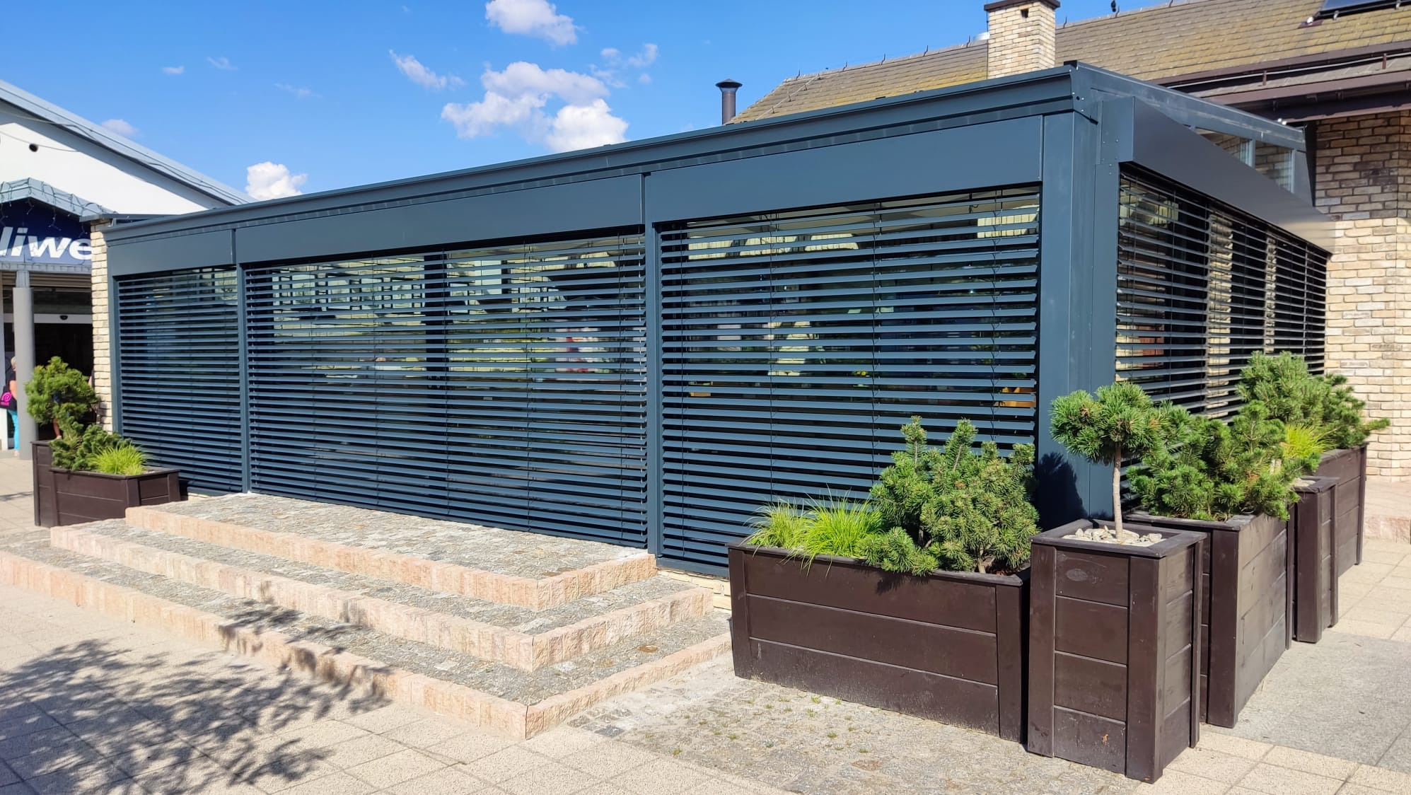 Zewnętrzne żaluzje fasadowe w kolorze antracytowym, zamontowane na budynku z cegły, z ozdobnymi roślinami w donicach. Widoczne schody wejściowe.