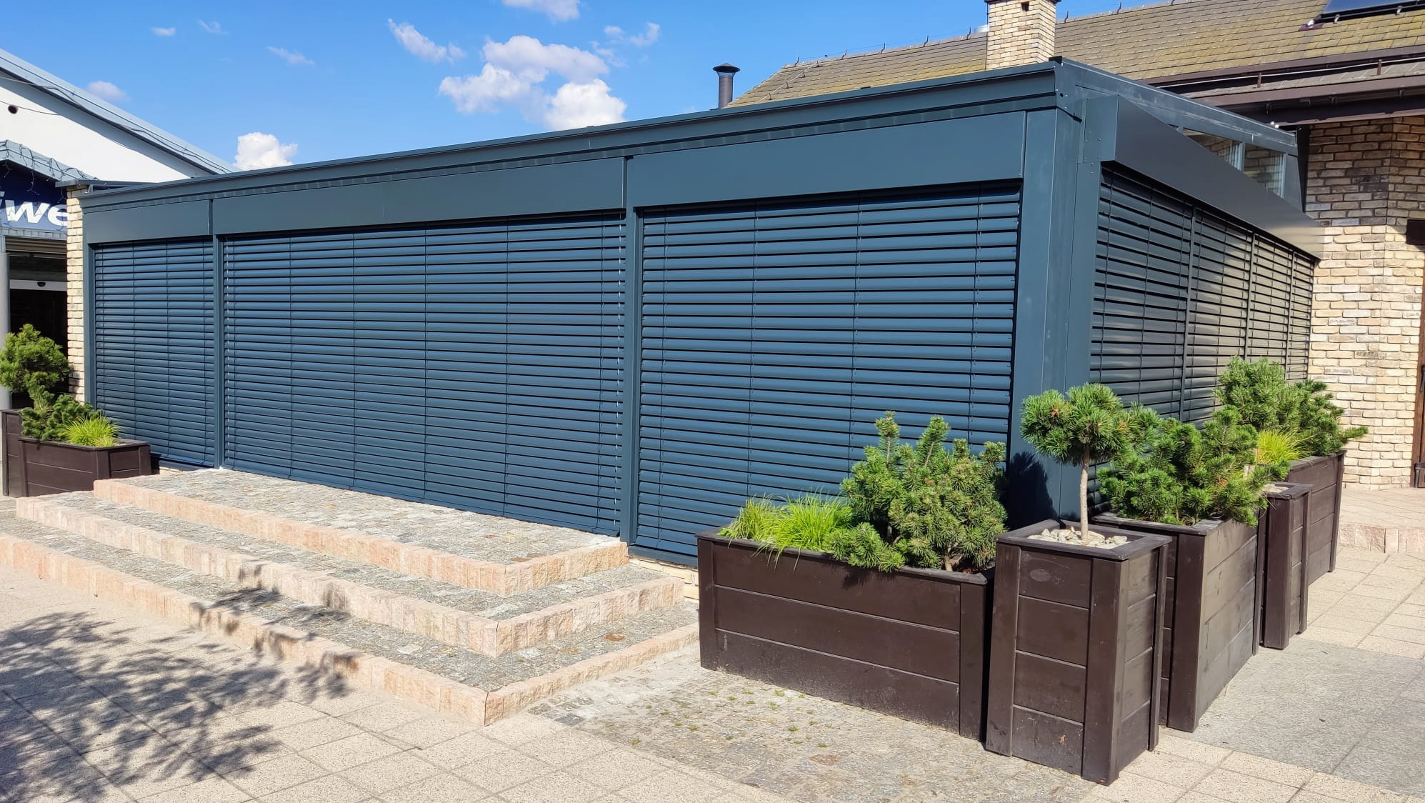 Zewnętrzne żaluzje fasadowe w kolorze grafitowym, zamontowane na budynku z cegły. Cztery donice z zielenią przed żaluzjami. Widoczne schody wejściowe.