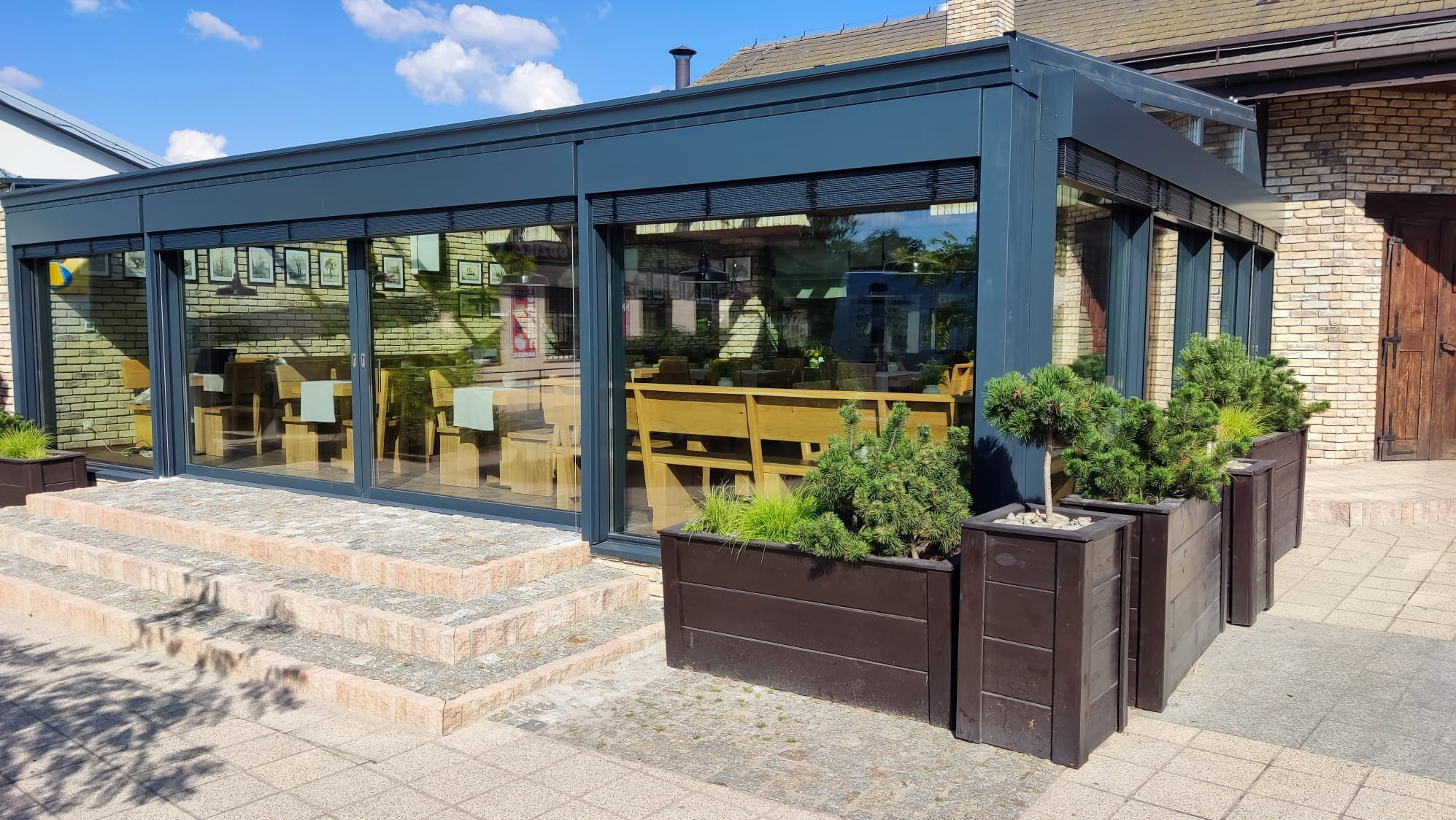 Zadaszony taras z aluminiową konstrukcją i przesuwnymi oknami, widok z zewnątrz. Dekoracyjne rośliny w donicach. Wnętrze widoczne przez okna.