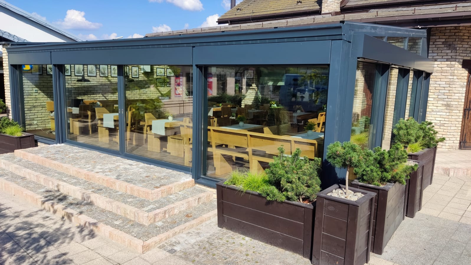 Szklana zabudowa tarasu restauracji w szarej ramie, z widocznymi stolikami i zielenią w donicach, na tle kamiennych schodów i chodnika.