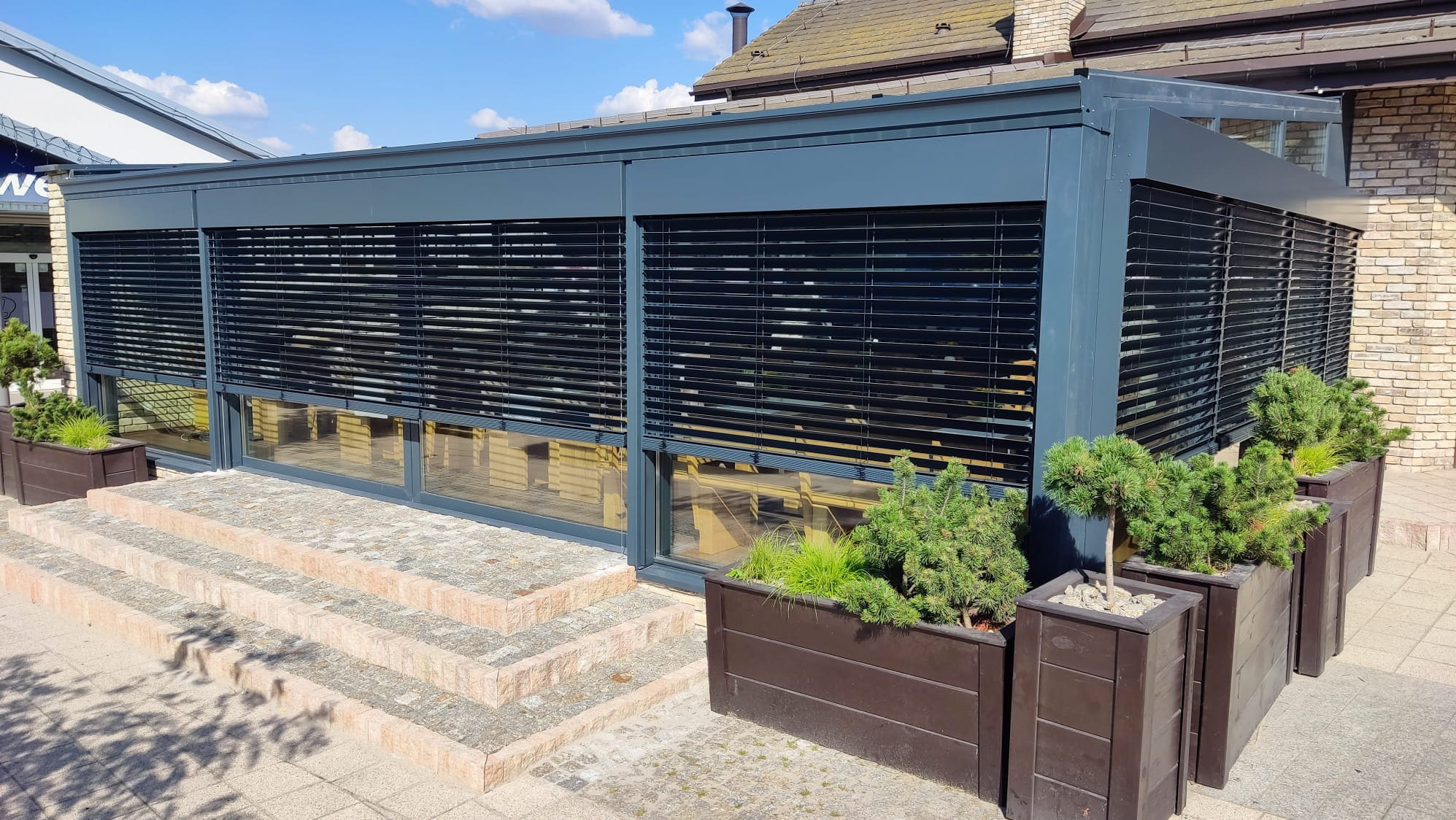 Nowoczesna zabudowa tarasowa z żaluzjami fasadowymi w kolorze grafitowym, zdobiona roślinami w donicach. Schody wejściowe z kostki brukowej.