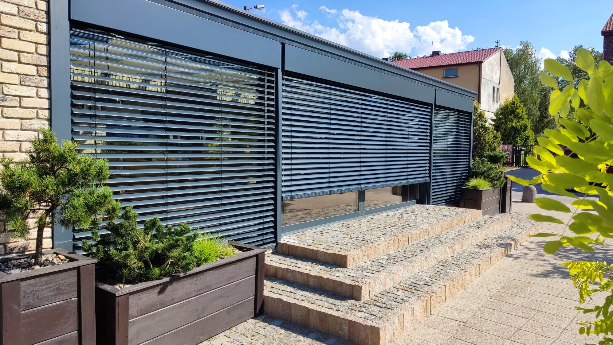 Elewacja budynku z szarymi żaluzjami fasadowymi, widoczne schody wejściowe i rośliny w donicach, błękitne niebo w tle.