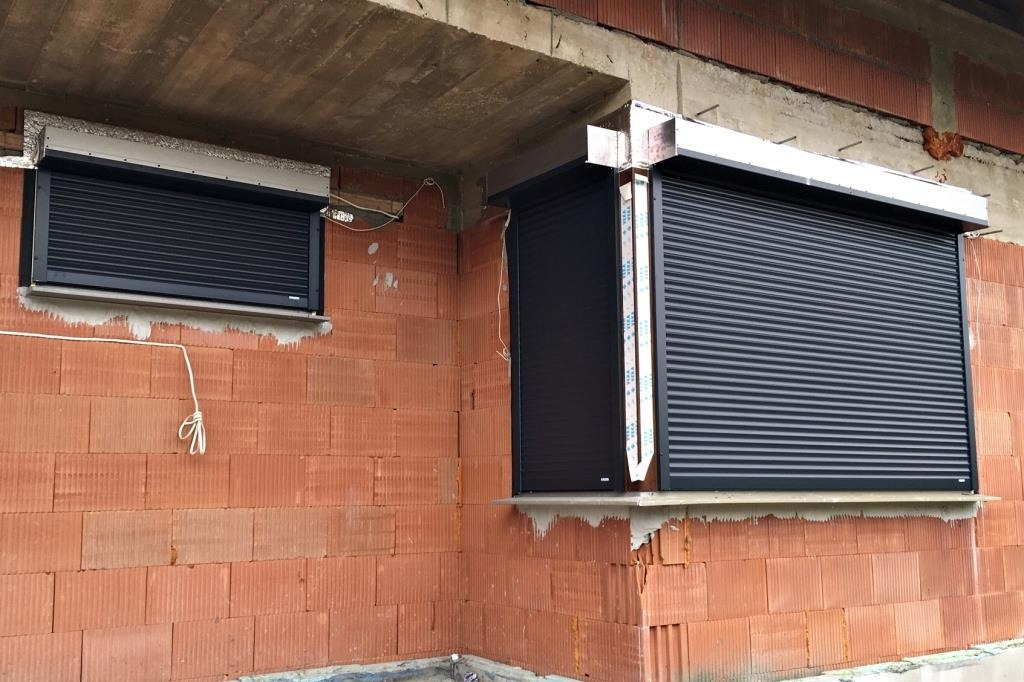 Ciemne rolety zamontowane w oknach budynku w trakcie budowy, widoczne cegły i surowy beton. Roleta po prawej stronie z listwami ochronnymi na boku.