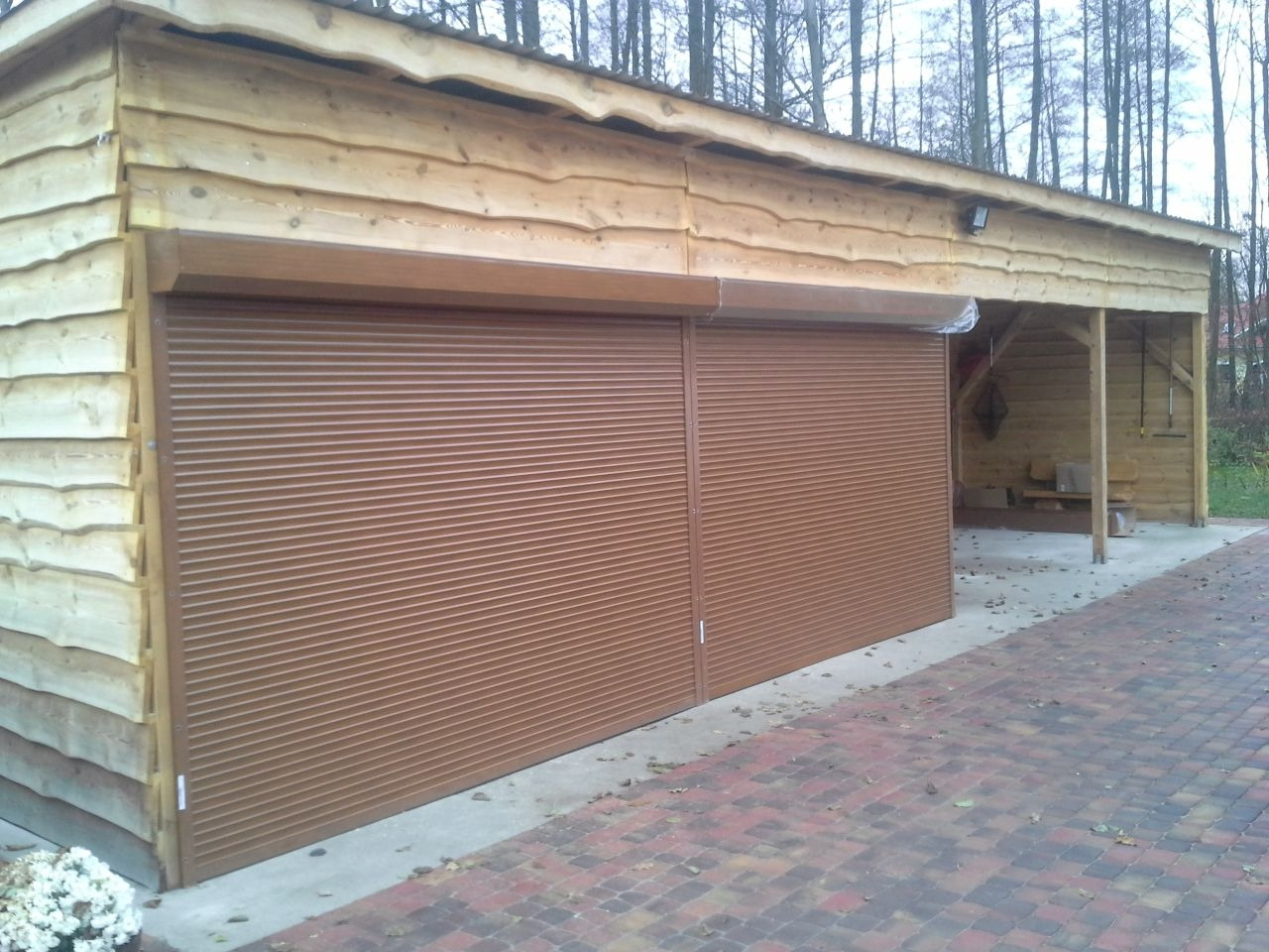 Brązowa roleta na drewnianym garażu z dobudowaną wiatą, widok na kostkę brukową i drzewa w tle. Zamknięta, pozioma roleta na garażu z desek.