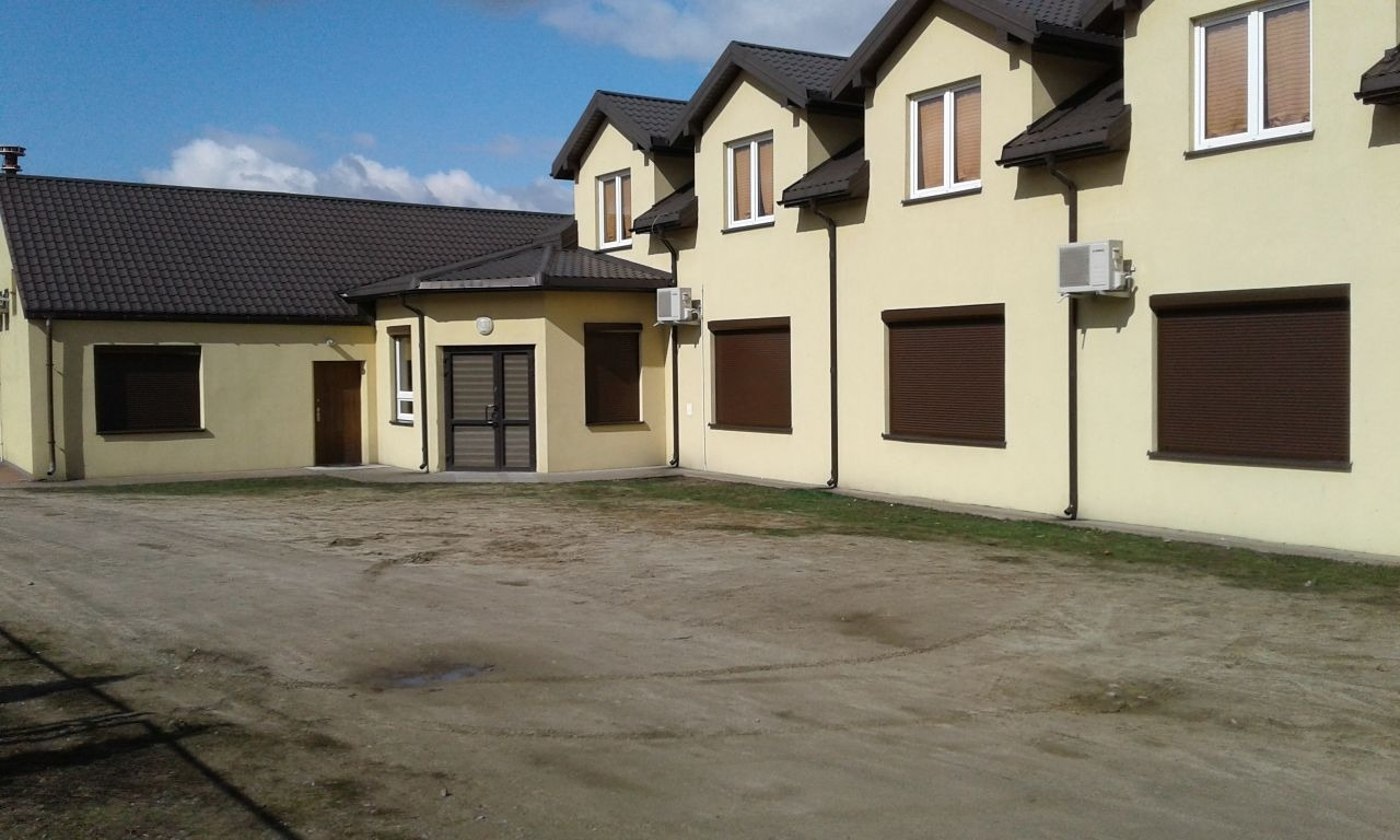 Żółty budynek z brązowymi roletami zewnętrznymi, widoczne klimatyzatory na ścianie. Ujęcie z zewnątrz, w tle błękitne niebo.