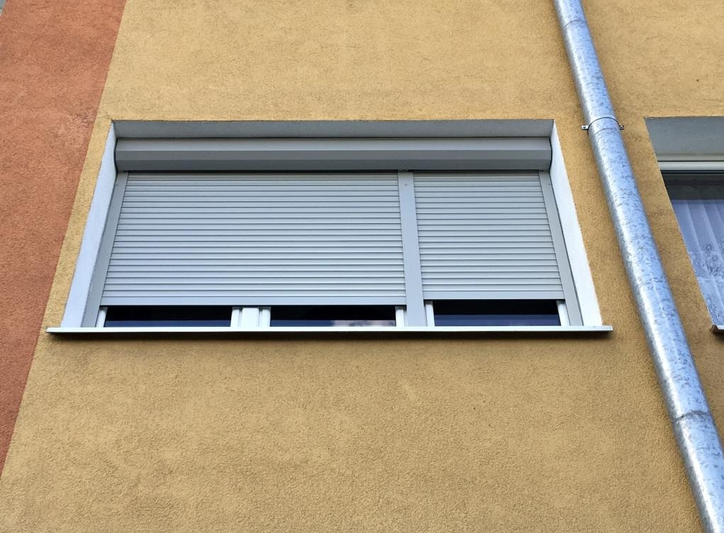 Zamknięta szara roleta zewnętrzna na oknie budynku o żółtej elewacji. Widoczna rynna. Detal architektoniczny, ochrona, nowoczesność.