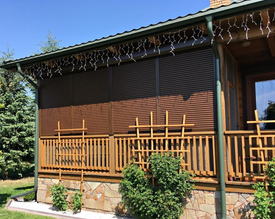 Zewnętrzne, brązowe rolety opuszczone na werandzie drewnianego domu z kamienną podmurówką, ozdobioną lampkami. Widok na ogród i błękitne niebo.