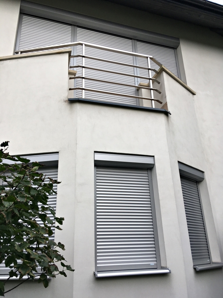 Elewacja budynku z oknami i balkonem, na których zamontowano szare rolety zewnętrzne z widocznym mechanizmem zwijania. Z lewej strony widoczny krzew.