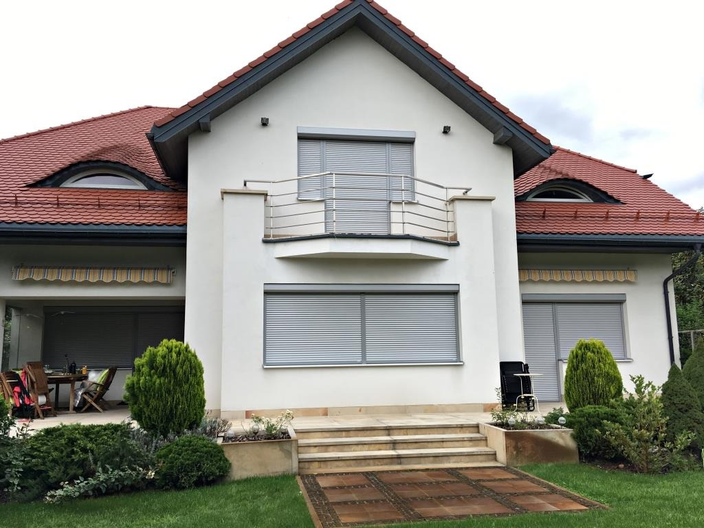 Elewacja domu z zamkniętymi, szarymi roletami zewnętrznymi na oknach i balkonie. Widoczny fragment tarasu i zadbany ogród.