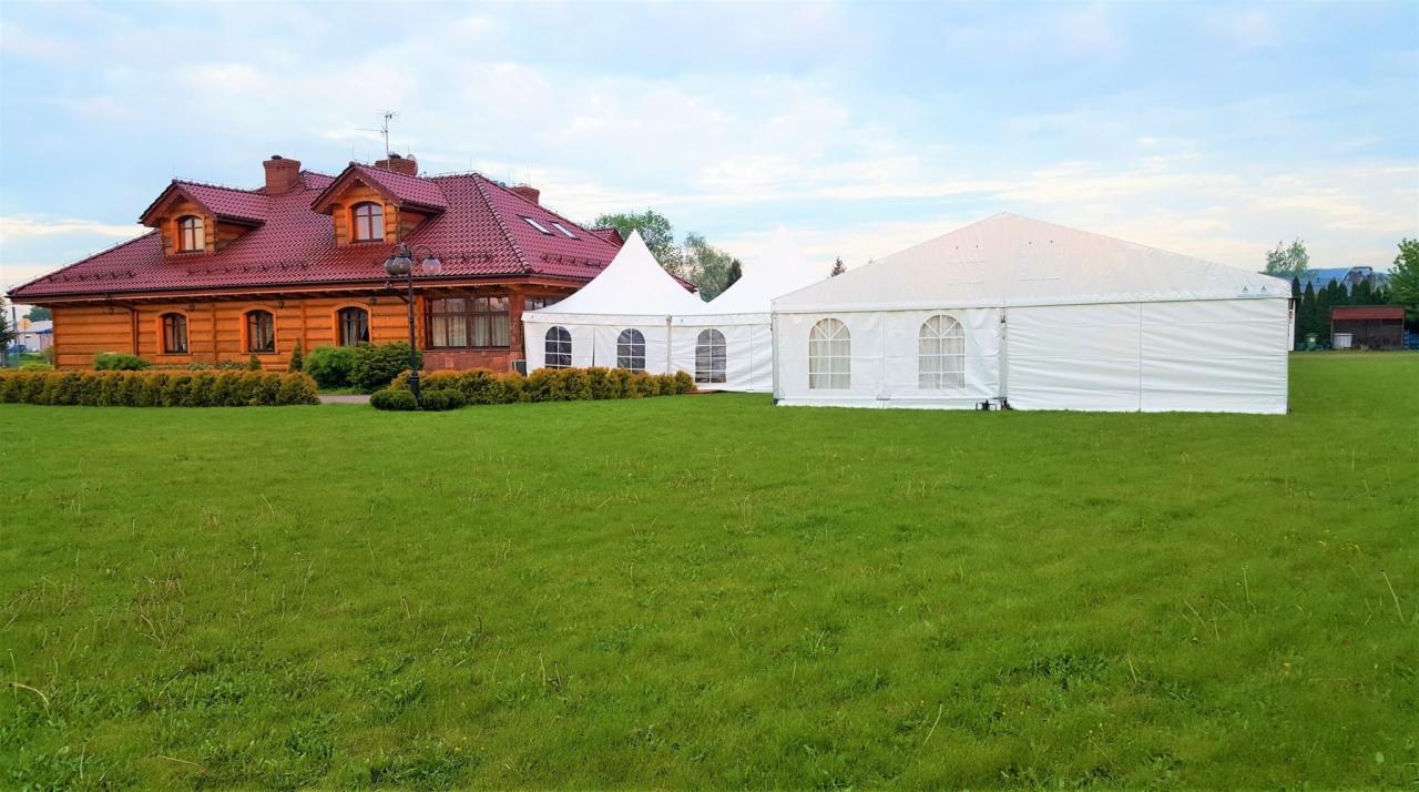 Rozstawione białe namioty imprezowe na zielonym trawniku, obok drewnianego domu z czerwonym dachem.