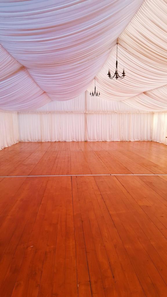 Pusta przestrzeń wewnątrz namiotu imprezowego z drewnianą podłogą i białym, drapowanym sufitem z wiszącymi żyrandolami.