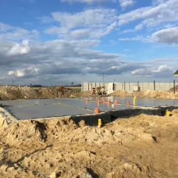 Wylany fundament pod dom z widoczną hydroizolacją, pomarańczowymi rurami instalacyjnymi i zarysami przyszłych ścian, na tle pochmurnego nieba.