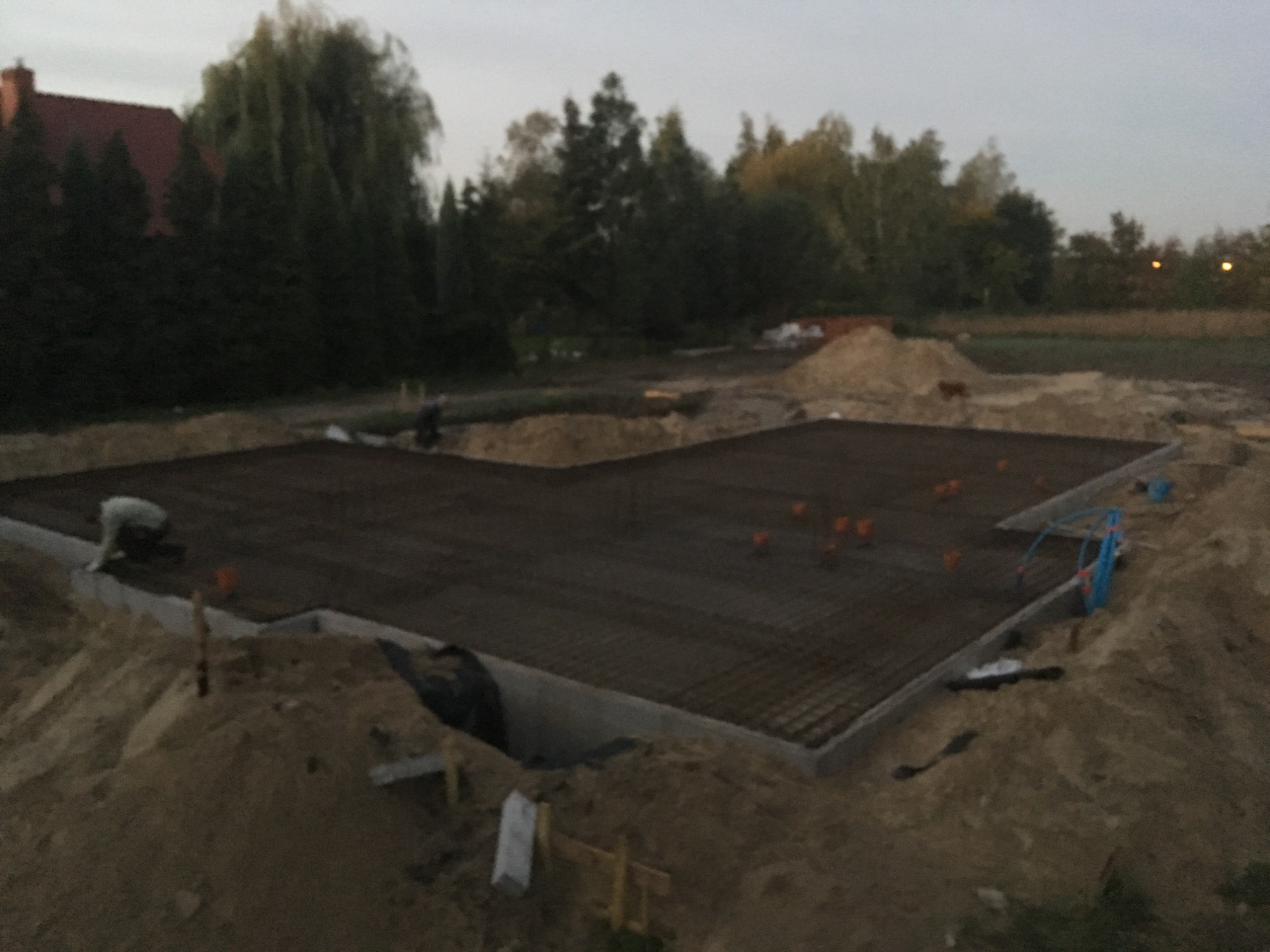 Przygotowany zbrojony fundament pod budynek w trakcie wylewania betonu, widoczne wystające rury instalacyjne i otaczający teren budowy z drzewami w tle.