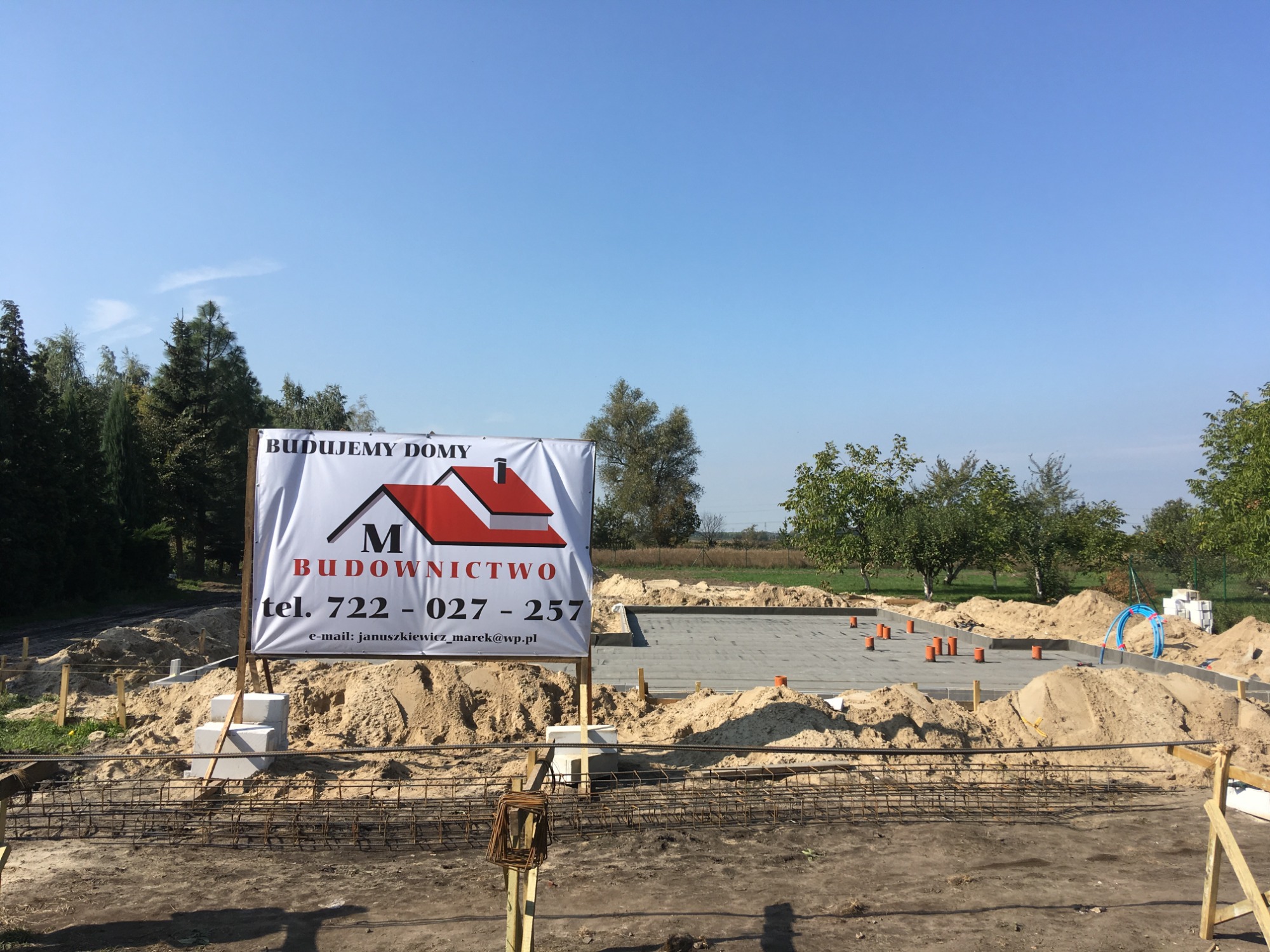 Fundamenty domu w trakcie budowy, widoczny zbrojony fundament, logo firmy budowlanej na tablicy reklamowej, teren budowy z piaskiem i narzędziami.