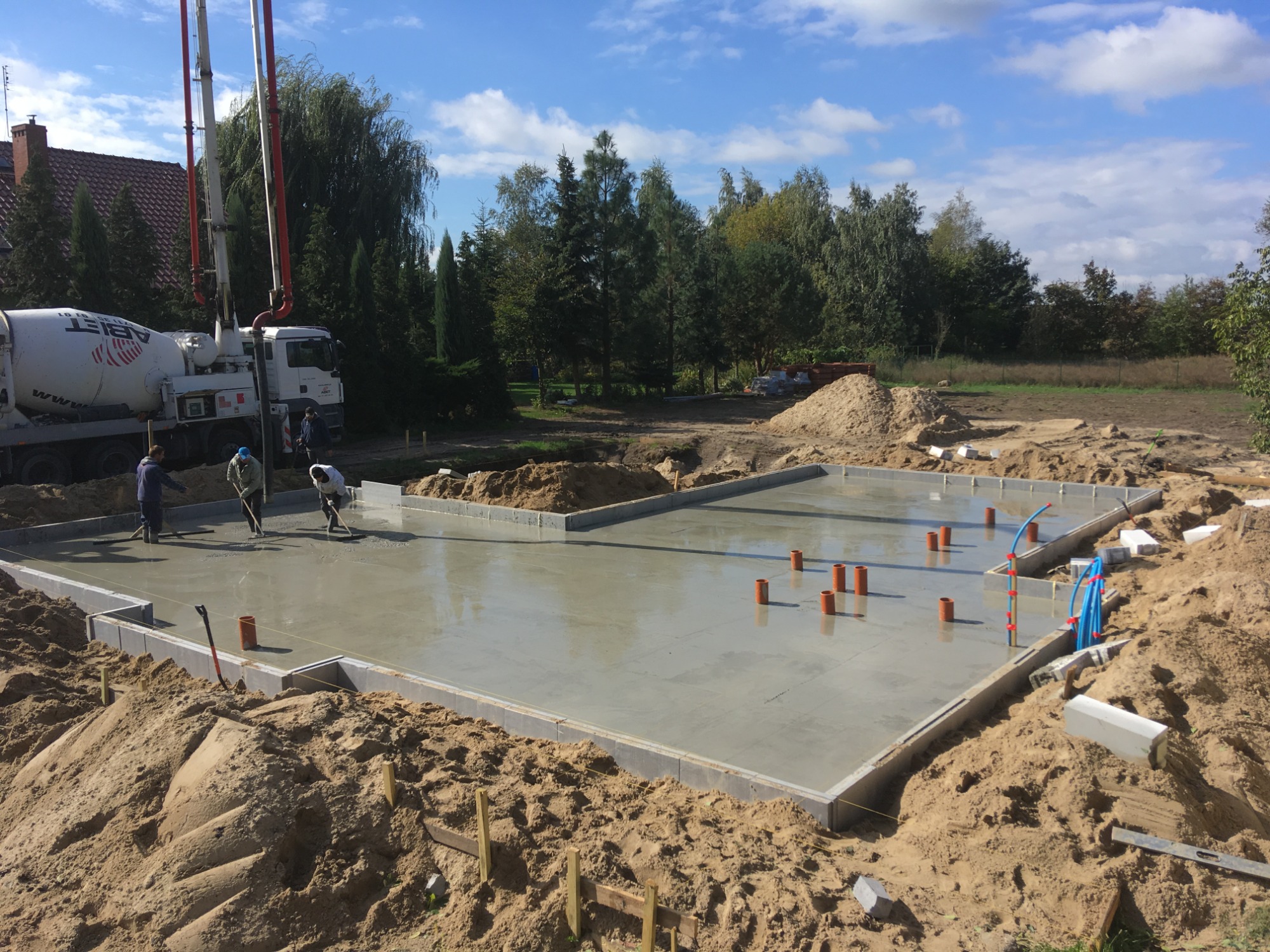 Wylewka fundamentowa z rurami instalacyjnymi, betonowany fundament domu jednorodzinnego, widoczny mikser z pompą do betonu i robotnicy zacierający beton, teren budowy w słoneczny dzień.