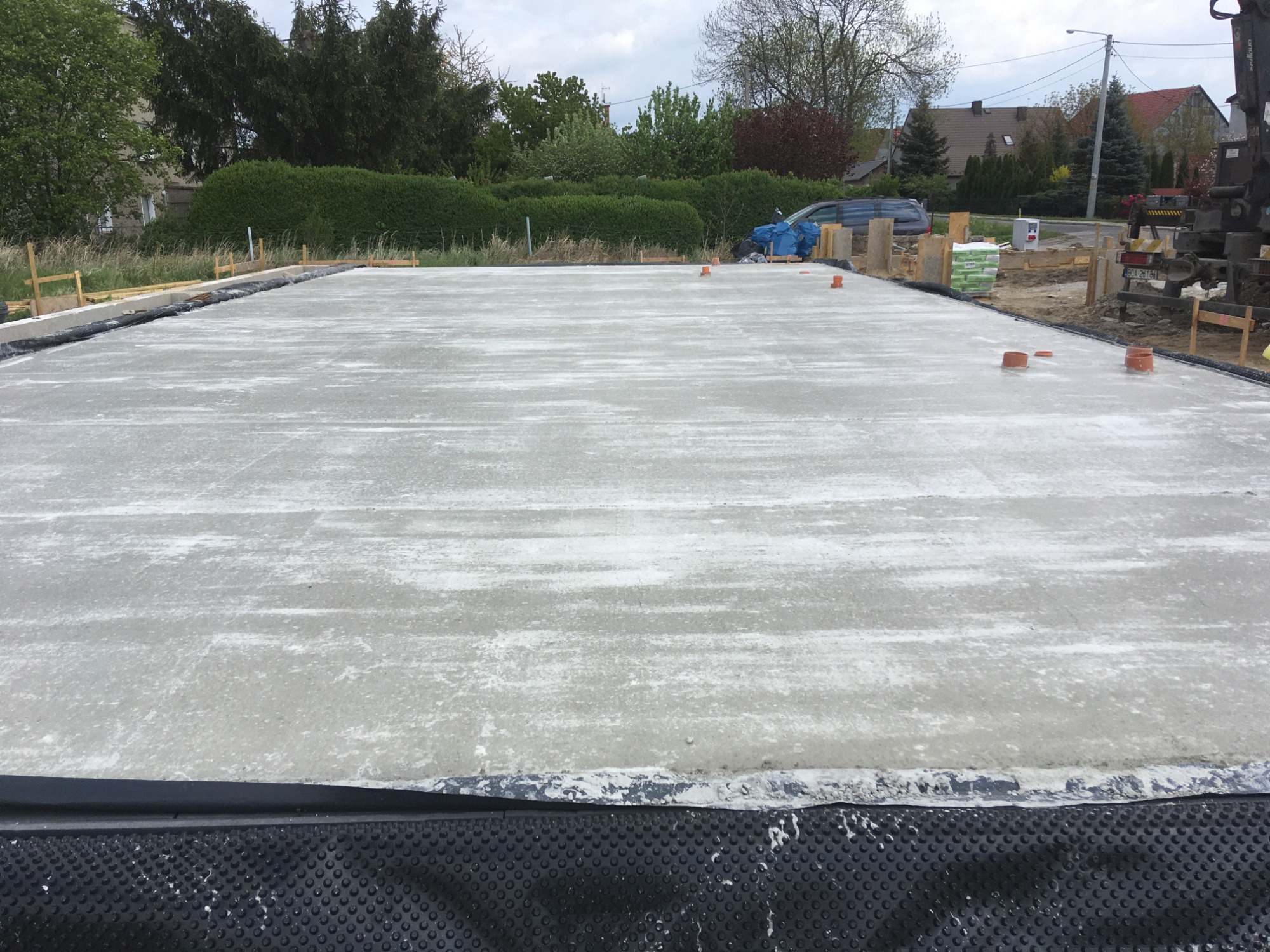 Wylana płyta fundamentowa z widoczną izolacją przeciwwilgociową, szalunkiem i wystającymi rurami kanalizacyjnymi, na tle zieleni i zabudowań mieszkalnych.