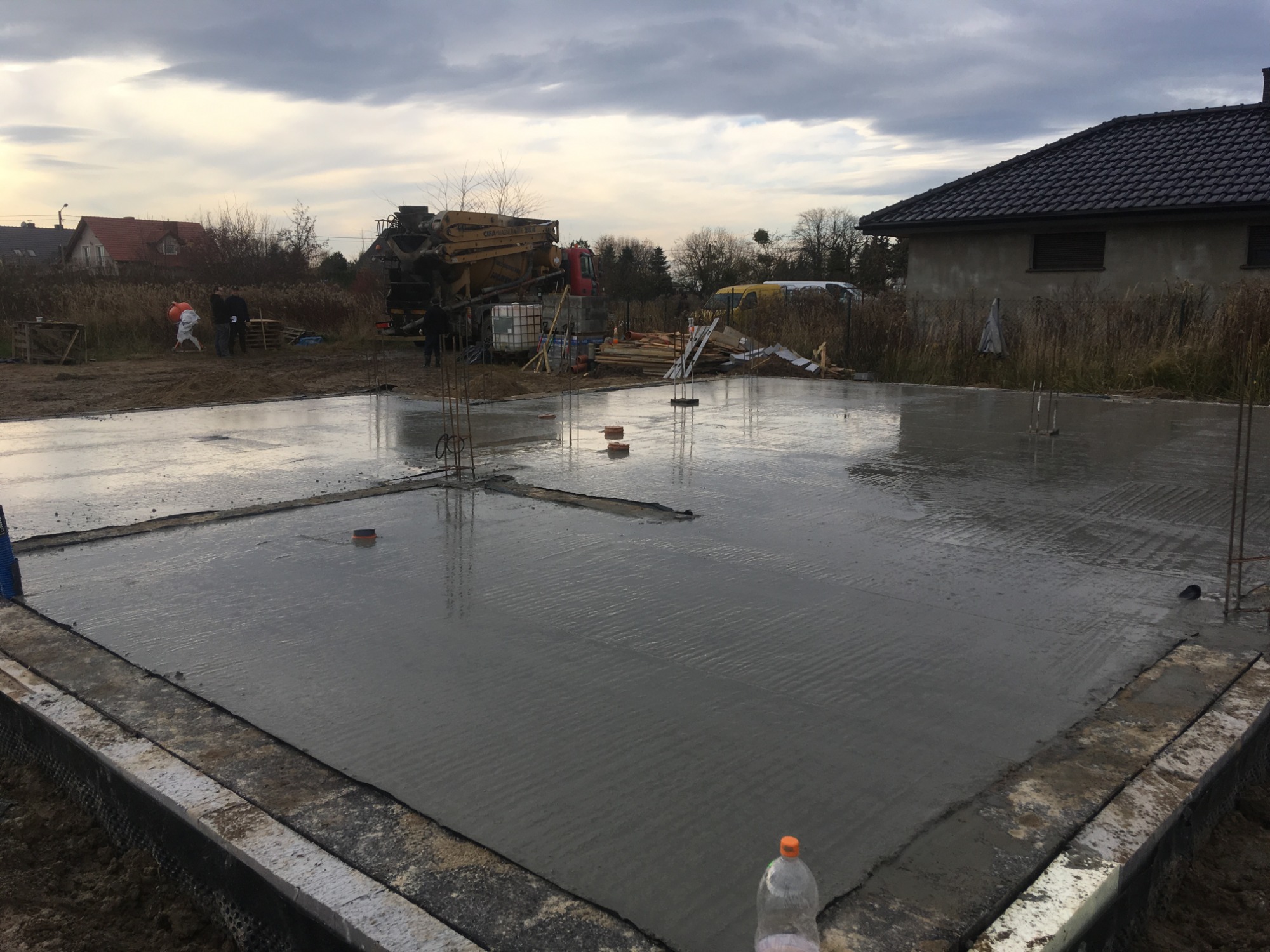 Wylany fundament pod budowę domu, widoczny beton, zbrojenia i przygotowane szalunki, w tle maszyna do betonu i ekipa budowlana.