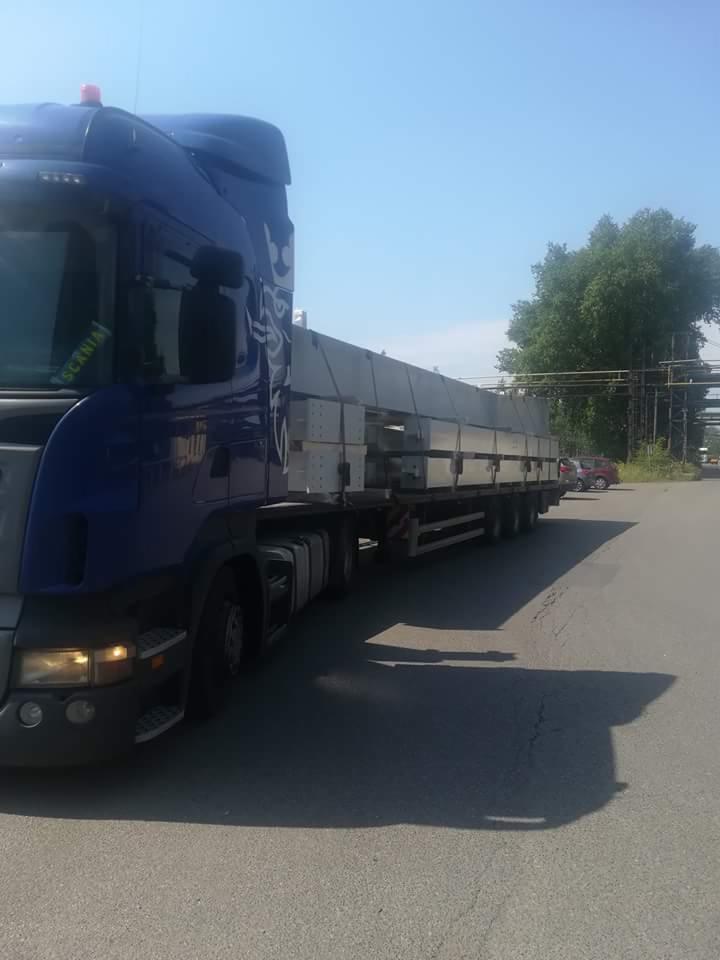 Niebieska ciężarówka Scania z ładunkiem betonowych prefabrykatów na naczepie, zaparkowana na asfaltowej drodze w słoneczny dzień.