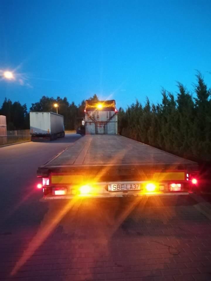 Płaska naczepa ciężarowa z widoczną tablicą rejestracyjną SBE LA37, zaparkowana na placu o zmierzchu, z innym pojazdem w tle.