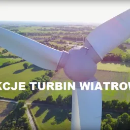 Biała turbina wiatrowa z widocznym dronem inspekcyjnym na tle zielonych pól i drzew.