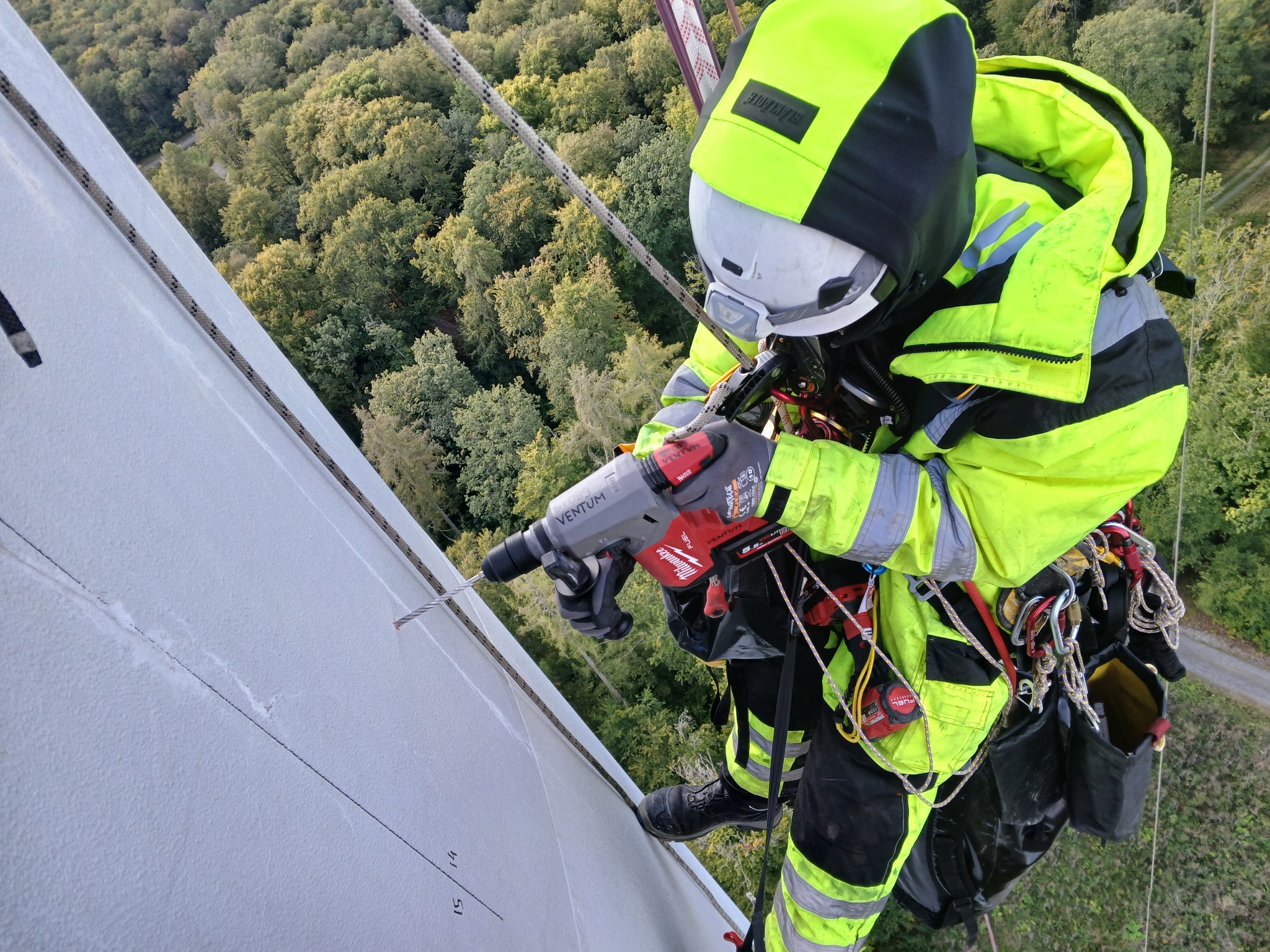 Pracownik w kasku i jaskrawej odzieży ochronnej, zabezpieczony linami, wierci w pionowej, białej konstrukcji na wysokości, z widokiem na drzewa w tle.