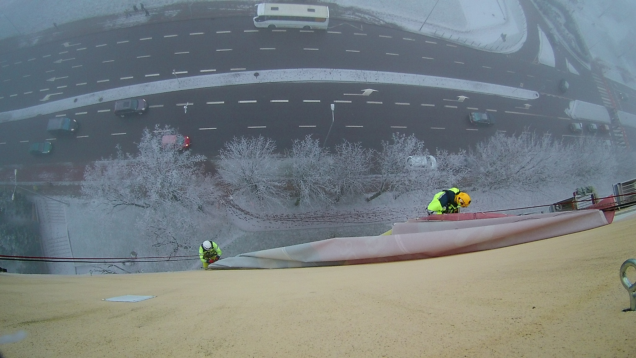 Dwóch pracowników w odblaskowych ubraniach zabezpiecza krawędź dachu budynku, widok z góry na zaśnieżone drzewa i ruch uliczny w pochmurny dzień.