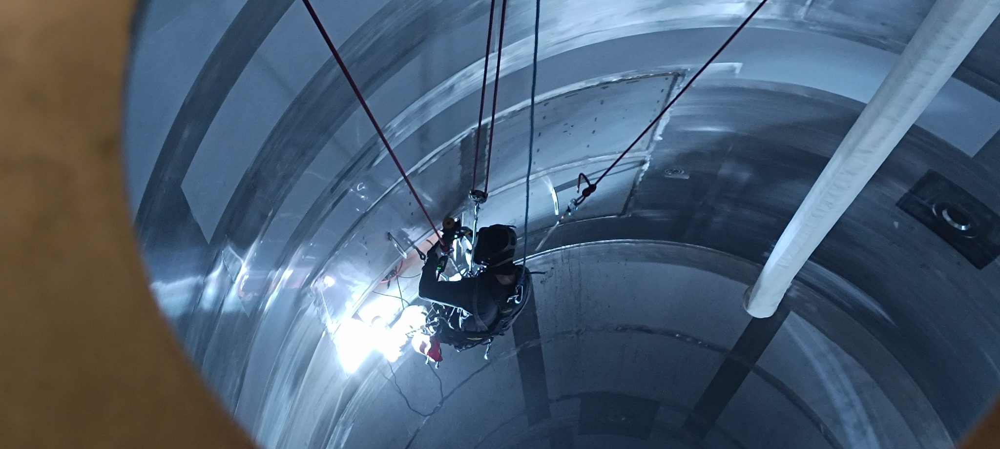 Pracownik w uprzęży, spawający metalową konstrukcję wewnątrz cylindrycznego zbiornika; widoczne iskry i liny asekuracyjne.