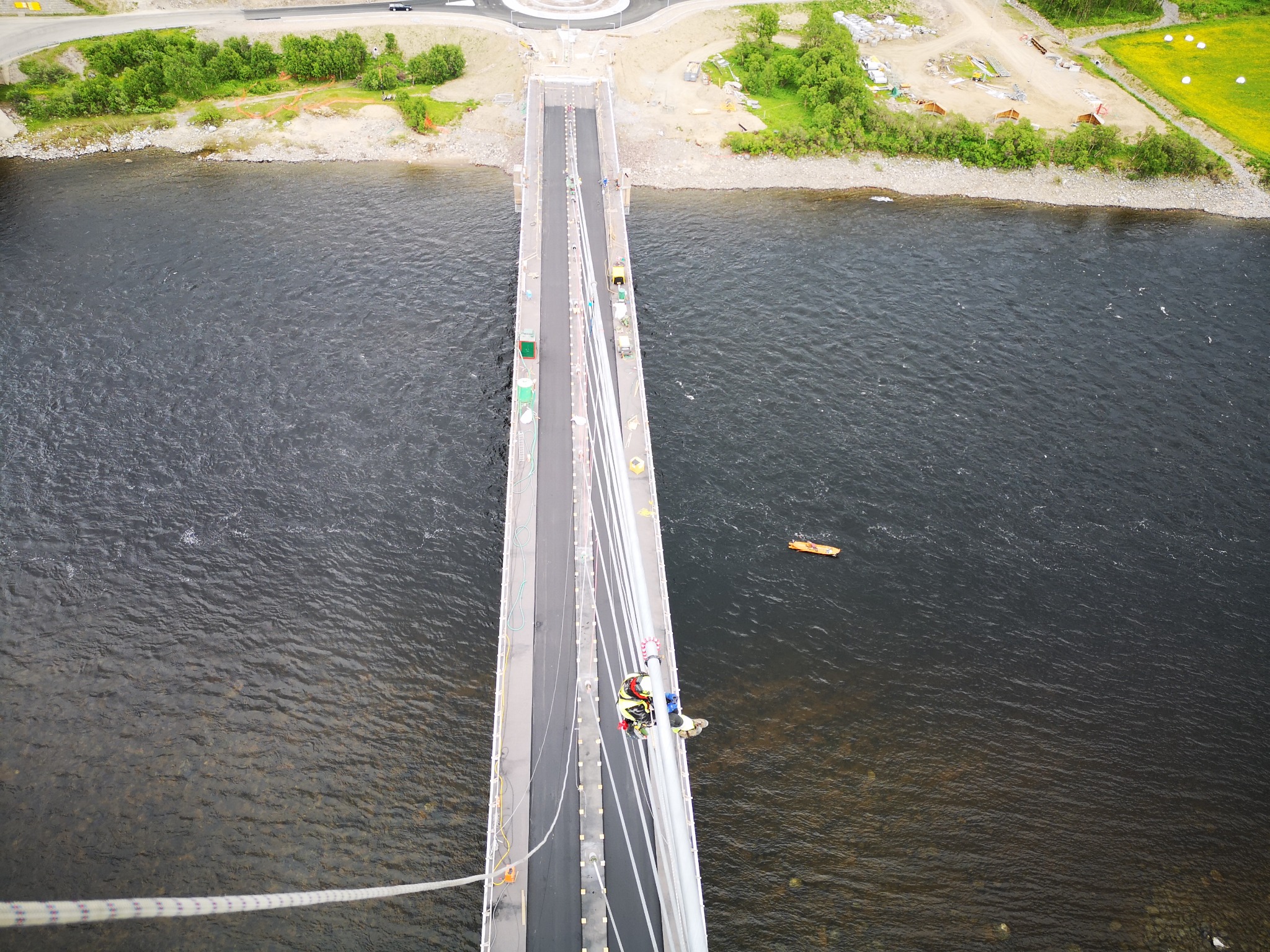 Pracownik w uprzęży, widok z góry, na pylonie mostu nad rzeką, podczas prac konserwacyjnych, z widocznym otoczeniem rzeki i zieleni.